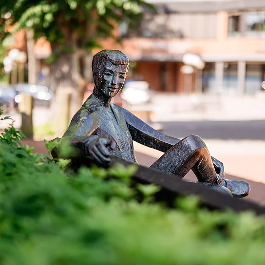 Bronzefigur mit einladender Handbewegung auf Bank