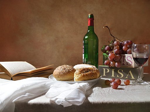 Tisch mit weißem Tischtuch, aufgeschlagenem Buch, Brot, Flasche Wein, Trauben und einem Weinglas